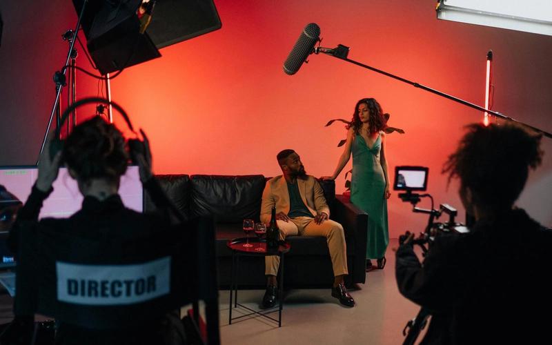 Set de gravação de comercial de TV com atores, diretor, refletores, microfone boom e câmera em estúdio profissional.