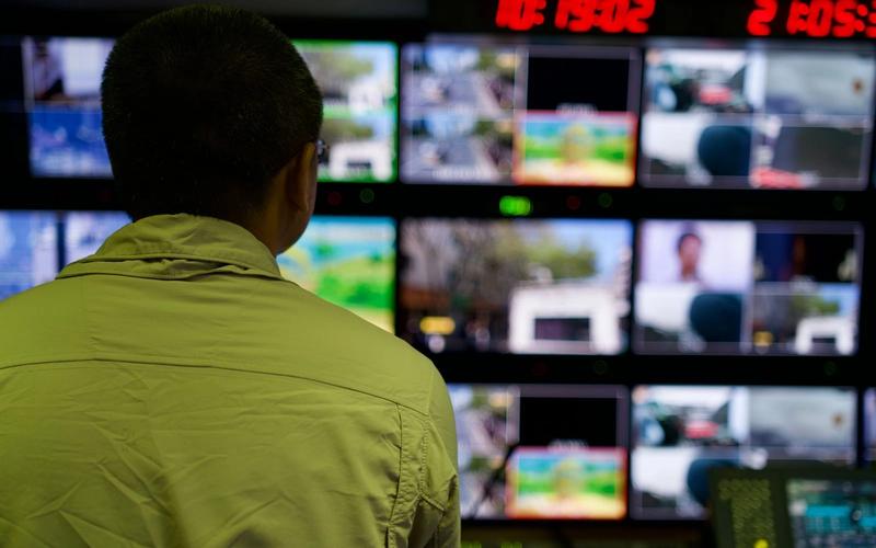 Profissional visto de costas observando um painel de monitores em uma sala de controle de transmissão ao vivo, com várias telas exibindo diferentes câmeras e um cronômetro digital vermelho ao fundo