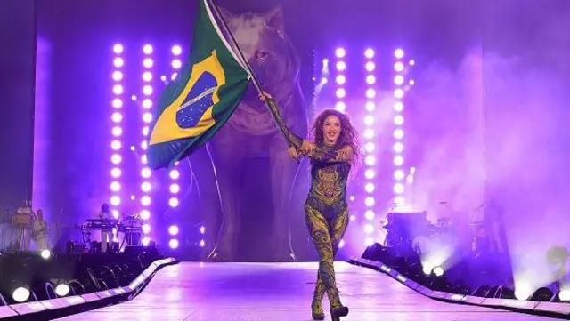 Shakira no show gratuito de copacabana no Rio de Janeiro, segurando uma bandeira do Brasil no palco com um lobo projetado atrás no cenário..