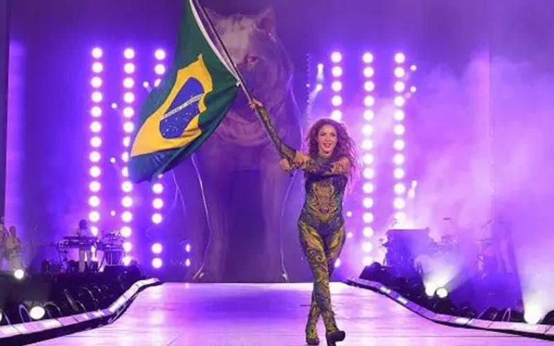 Shakira no show gratuito de copacabana no Rio de Janeiro, segurando uma bandeira do Brasil no palco com um lobo projetado atrás no cenário..