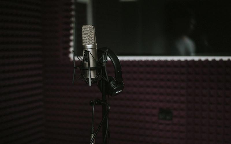 Microfone condensador profissional com fones de ouvido dentro de uma cabine de gravação com isolamento acústico, e a silhueta de uma pessoa visível atrás do vidro do estúdio de dublagem
