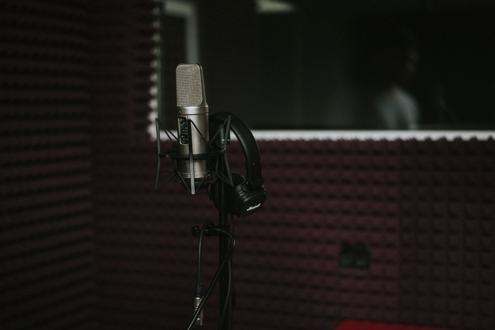 Microfone condensador profissional com fones de ouvido dentro de uma cabine de gravação com isolamento acústico, e a silhueta de uma pessoa visível atrás do vidro do estúdio de dublagem