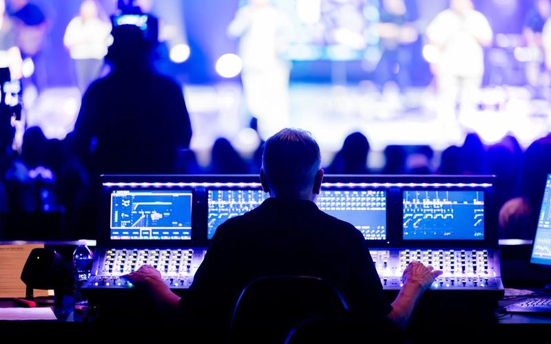 Técnico de som visto de costas operando uma mesa de mixagem digital durante um show ao vivo, com telas exibindo curvas de equalização e o palco iluminado em azul e roxo ao fundo