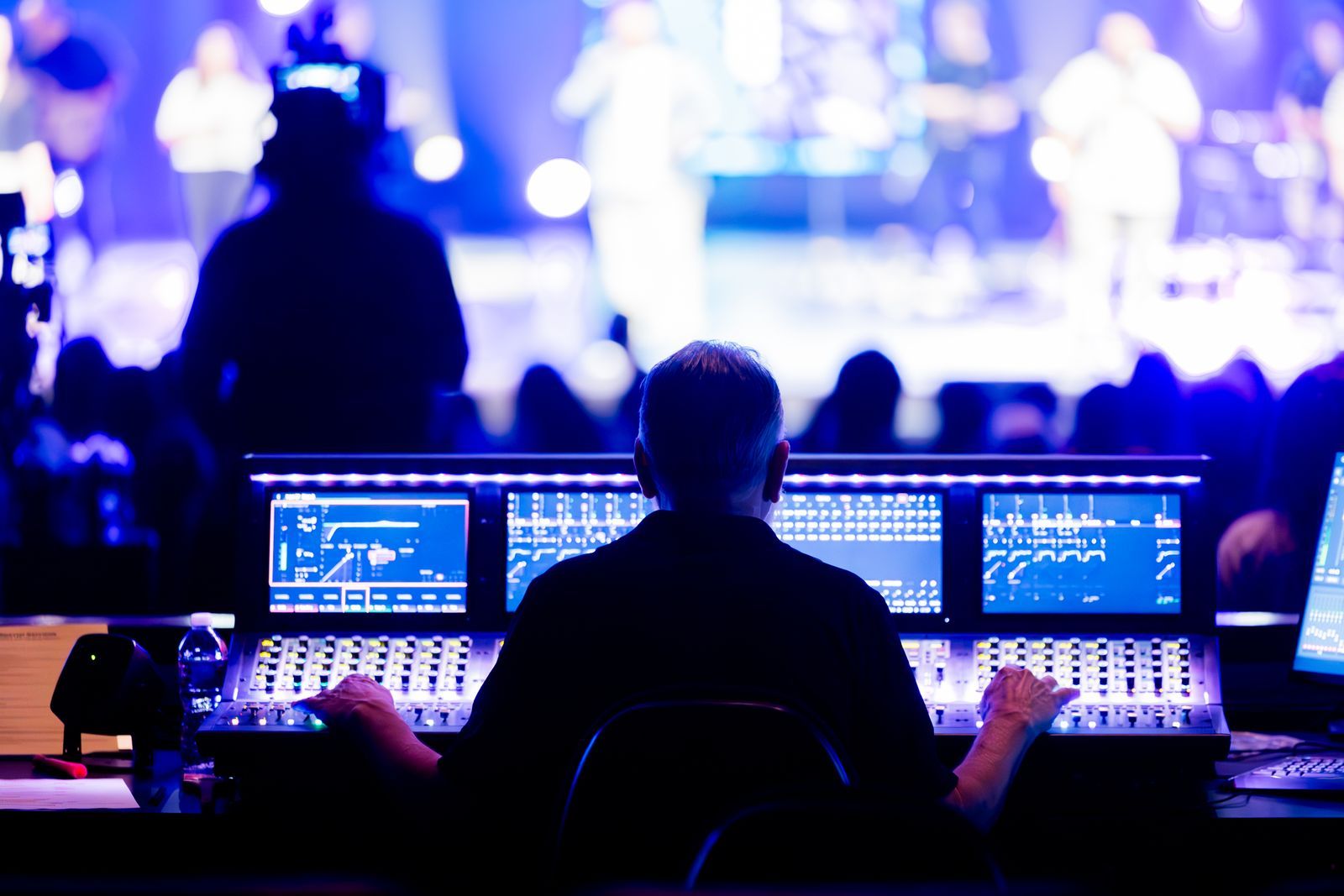 Técnico de som visto de costas operando uma mesa de mixagem digital durante um show ao vivo, com telas exibindo curvas de equalização e o palco iluminado em azul e roxo ao fundo