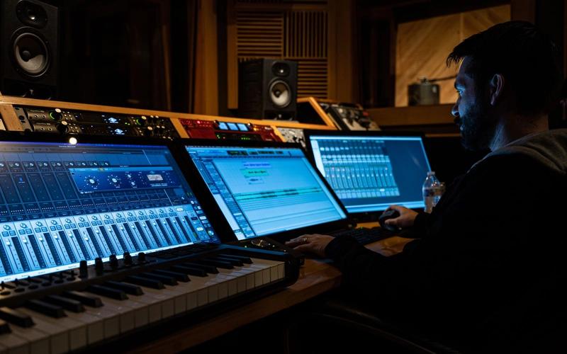 Compositor trabalhando em estúdio de gravação com teclado MIDI, mesa de mixagem e telas de software de áudio, ambiente cinematográfico e iluminação baixa, sugerindo a produção de uma trilha sonora