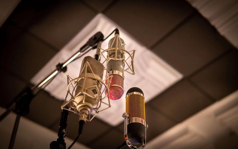 Três microfones de estúdio diferentes em suportes anti-choque, vistos de baixo para cima dentro de uma sala de gravação com tratamento acústico no teto.