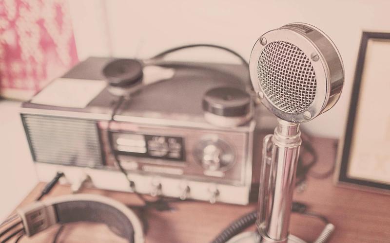 Microfone cromado antigo ao lado de um rádio receptor clássico, em tom sépia nostálgico, evocando a era de ouro da locução.