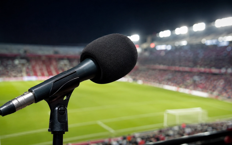 Microfone de transmissão esportiva em primeiro plano, com gramado e arquibancada lotada de um estádio à noite ao fundo, representando a cobertura jornalística da Copa do Mundo 2026