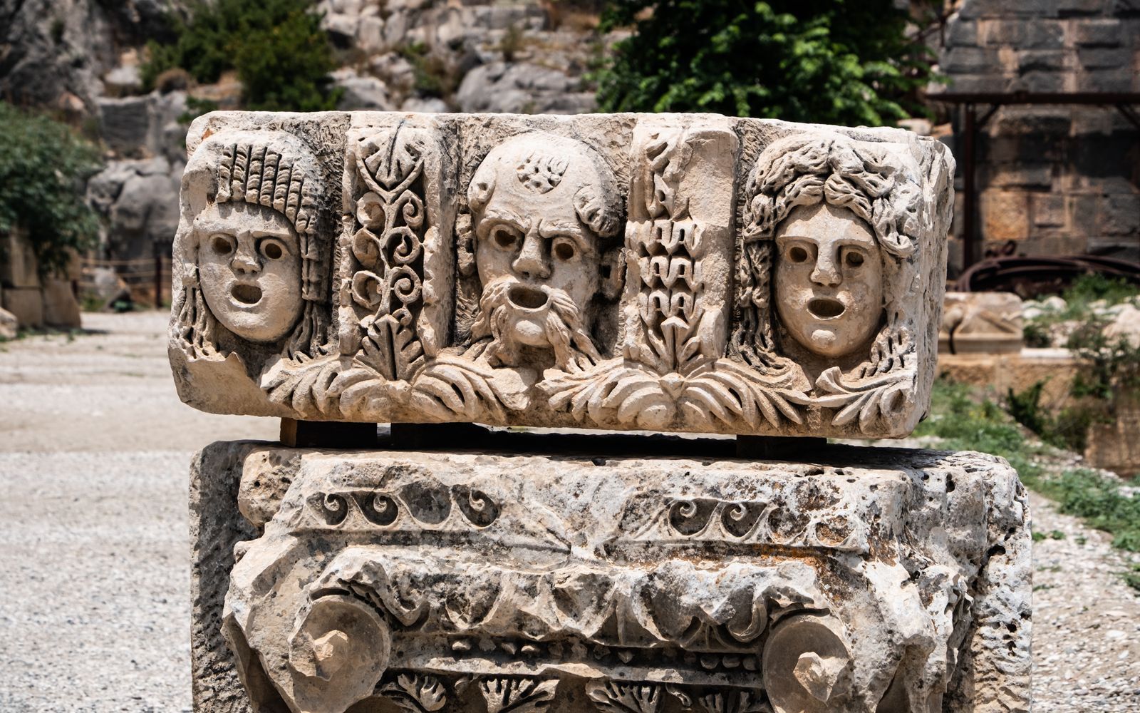 Relevo de pedra antigo com três máscaras gregas de teatro esculpidas lado a lado, em sítio arqueológico ao ar livre