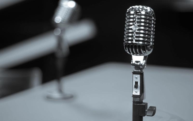 Microfone retrô cromado sobre pedestal em estúdio, em tons de preto e branco, evocando a era de ouro do rádio e da narração esportiva
