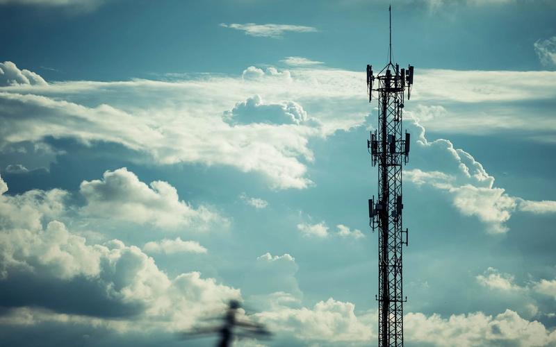 Torre de telecomunicações silhuetada contra um céu dramático de nuvens, representando a infraestrutura que transmite alertas de emergência para celulares