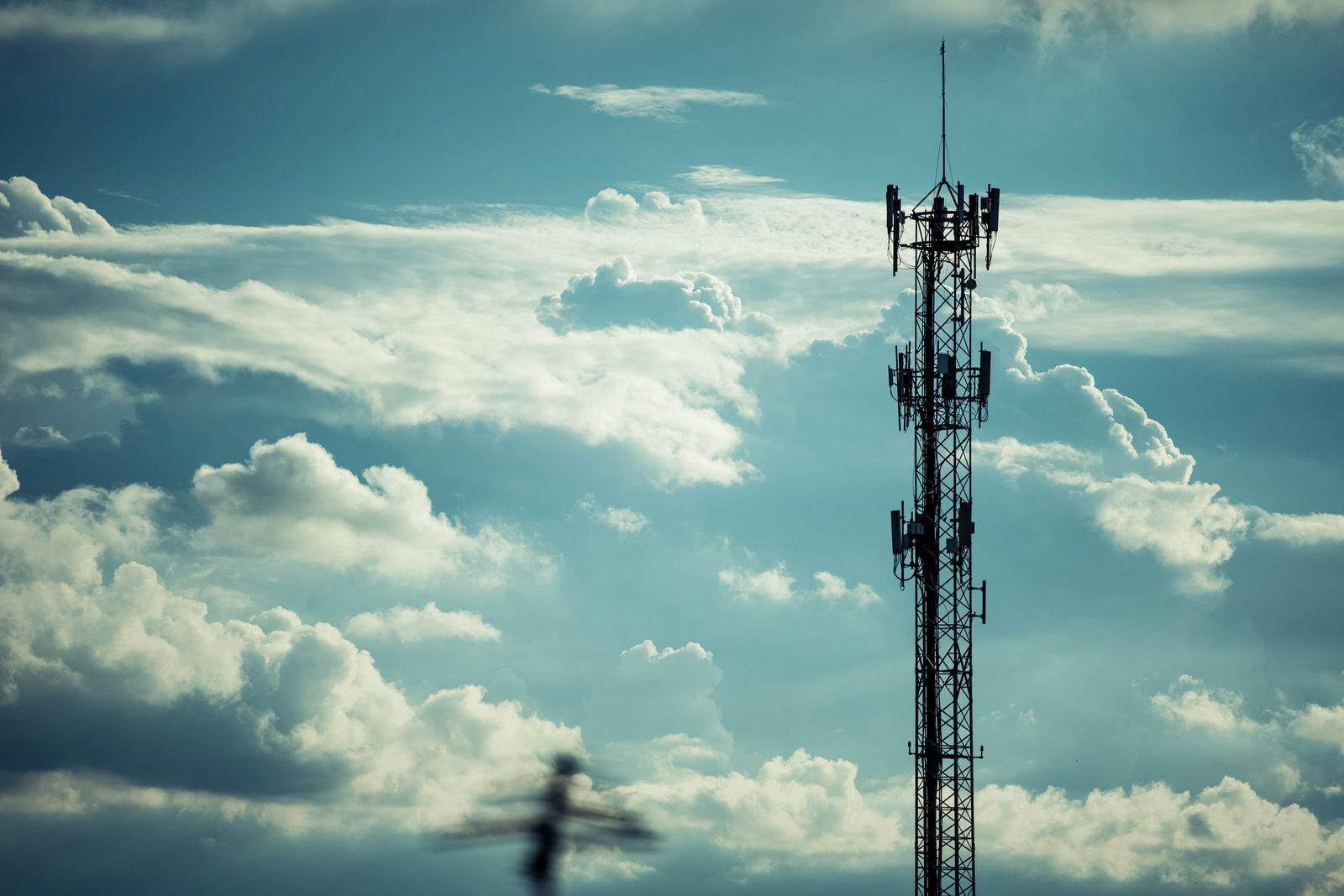 Torre de telecomunicações silhuetada contra um céu dramático de nuvens, representando a infraestrutura que transmite alertas de emergência para celulares