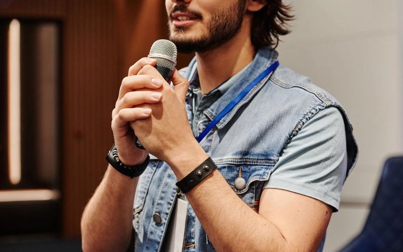 Homem de colete jeans falando ao microfone durante uma apresentação para uma plateia.