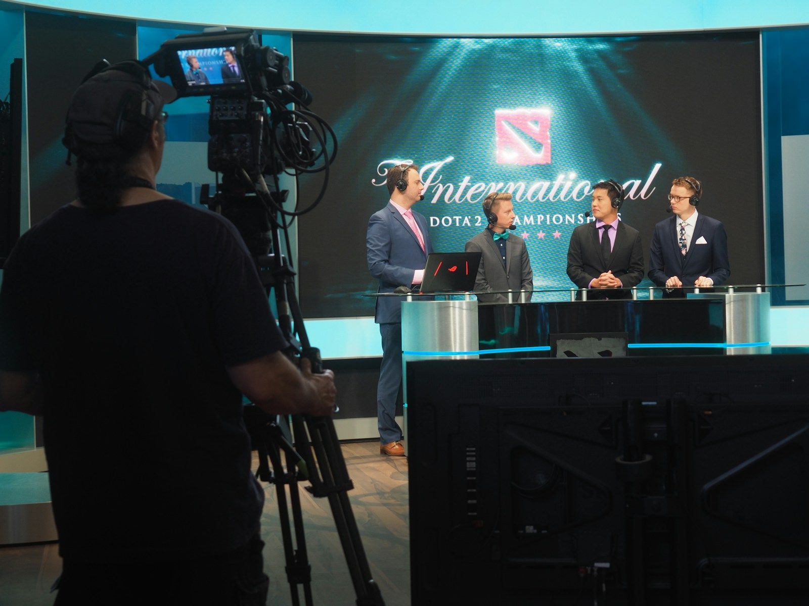 Três homens de terno sendo filmados numa mesa de transmissão de um campeonato de e-sports, com câmeras profissionais ao redor