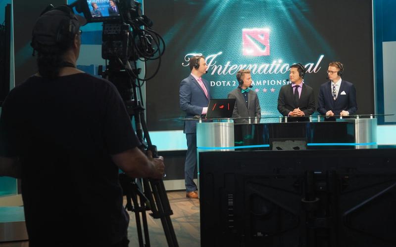 Três homens de terno sendo filmados numa mesa de transmissão de um campeonato de e-sports, com câmeras profissionais ao redor