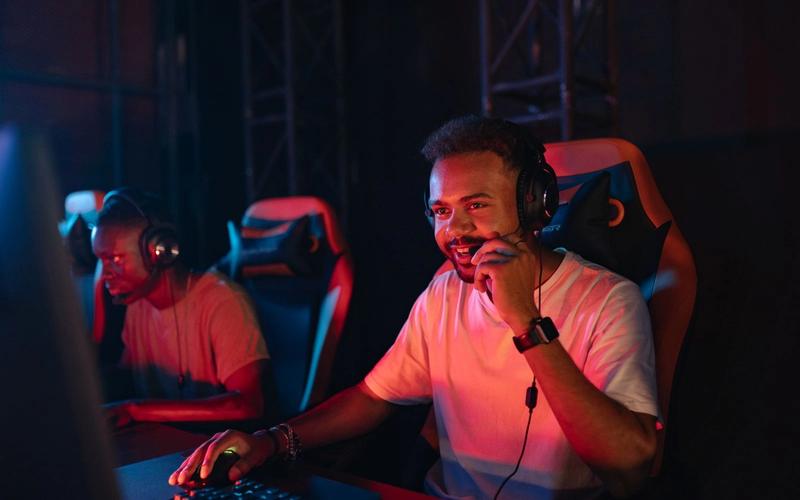 Dois jovens jogadores concentrados, de fones de ouvido com microfone, em um ambiente gamer com iluminação vermelha e azul.