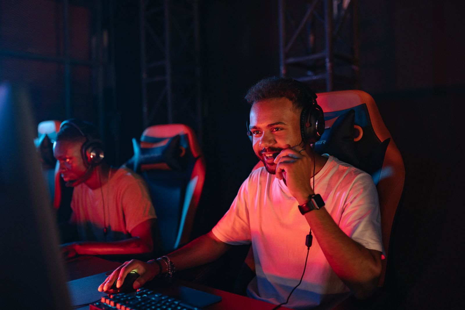 Dois jovens jogadores concentrados, de fones de ouvido com microfone, em um ambiente gamer com iluminação vermelha e azul.