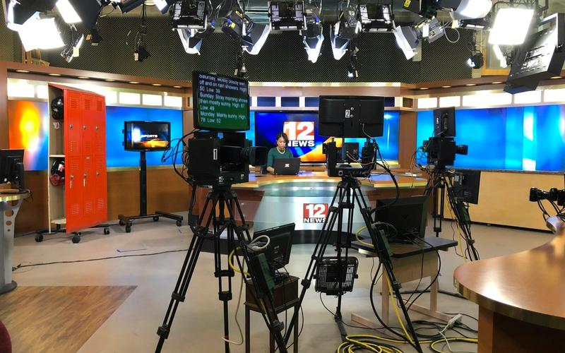 Estúdio de televisão profissional com câmeras, iluminação e bancada de telejornal, representando o curso de Apresentação de TV e Mídias da Escola de Rádio.