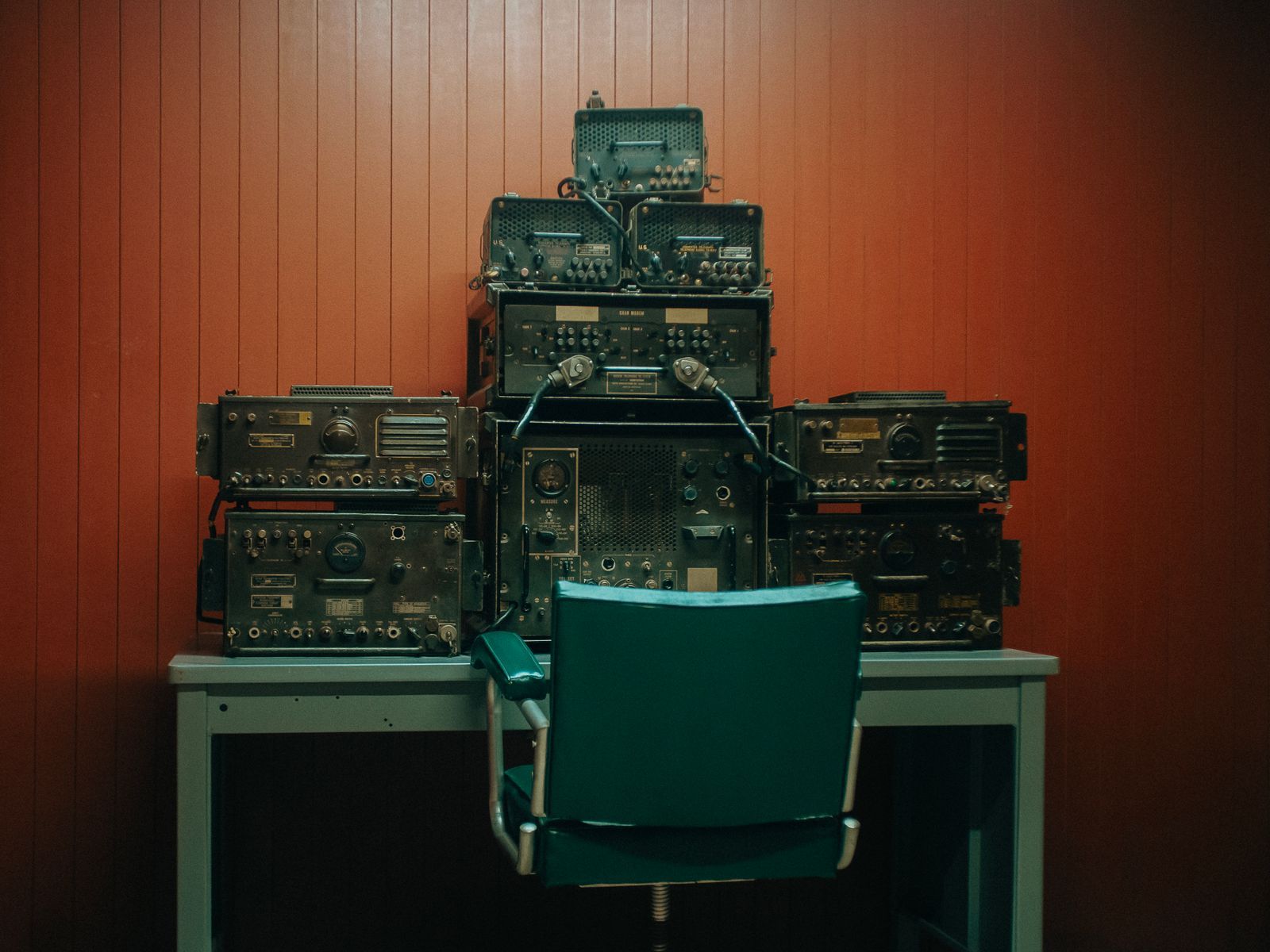 Estação de rádio vintage com equipamentos empilhados contra parede de madeira laqueada em tom terracota, cadeira de escritório retrô verde em primeiro plano