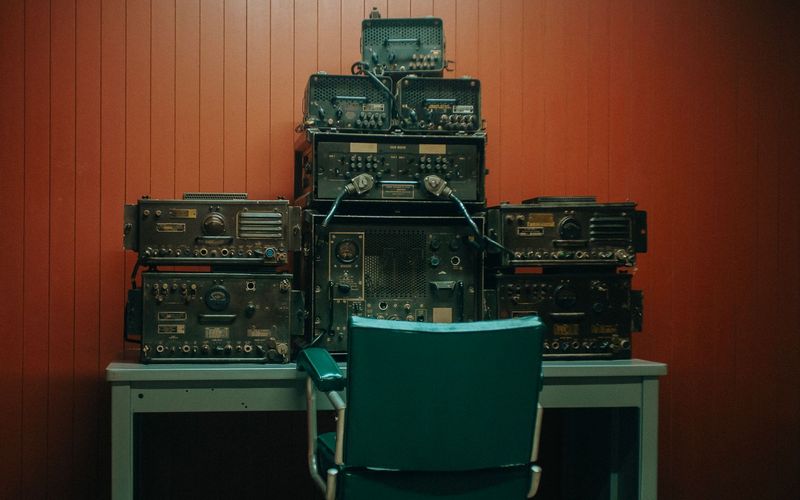 Estação de rádio vintage com equipamentos empilhados contra parede de madeira laqueada em tom terracota, cadeira de escritório retrô verde em primeiro plano