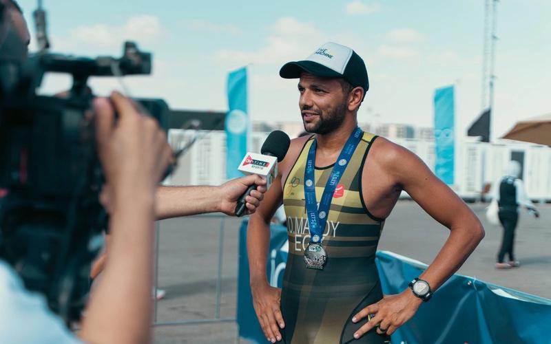 Repórter de campo entrevista um atleta logo após uma prova, segurando o microfone enquanto um cinegrafista grava ao ar livre