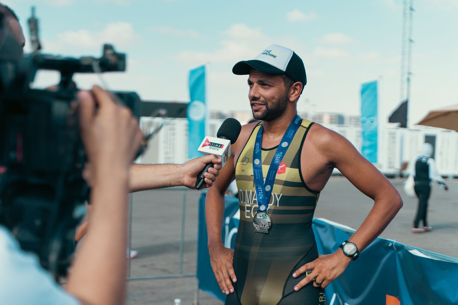 Repórter de campo entrevista um atleta logo após uma prova, segurando o microfone enquanto um cinegrafista grava ao ar livre