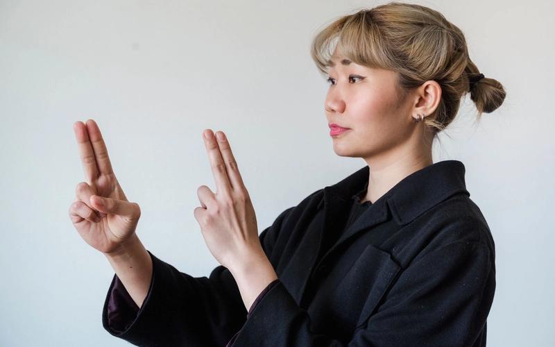 Mulher de perfil, em pé contra um fundo branco, fazendo um sinal de Libras com as duas mãos levantadas, dedos indicador e médio estendidos, vestindo um casaco preto.