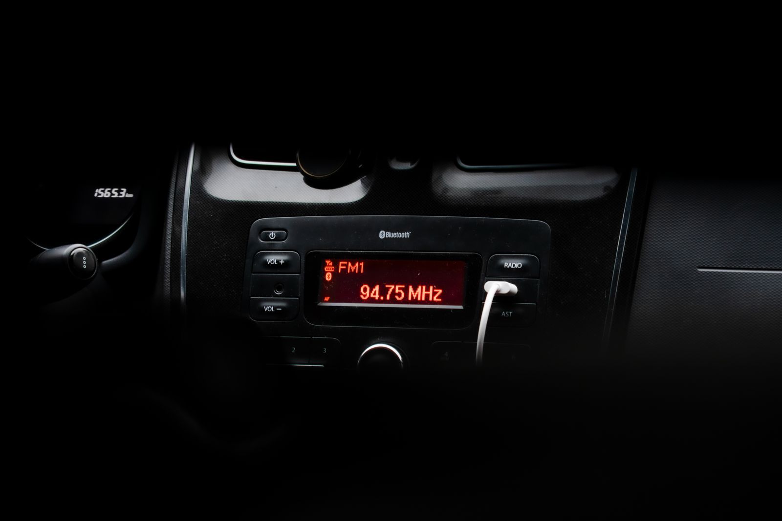 Close-up do painel escuro de um carro à noite, mostrando o display vermelho do rádio sintonizado em FM1 94.75 MHz, com o volante desfocado ao fundo