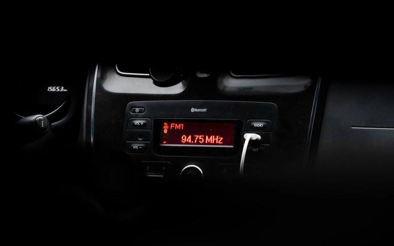 Close-up do painel escuro de um carro à noite, mostrando o display vermelho do rádio sintonizado em FM1 94.75 MHz, com o volante desfocado ao fundo