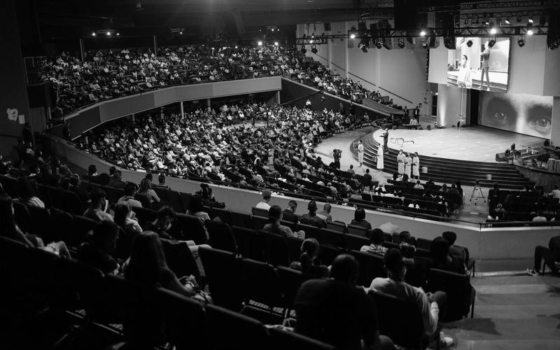 Foto em preto e branco de um auditório lotado visto do alto, com a plateia nas arquibancadas voltada para o palco iluminado e um grande telão ao fundo, evocando a era dos programas de auditório