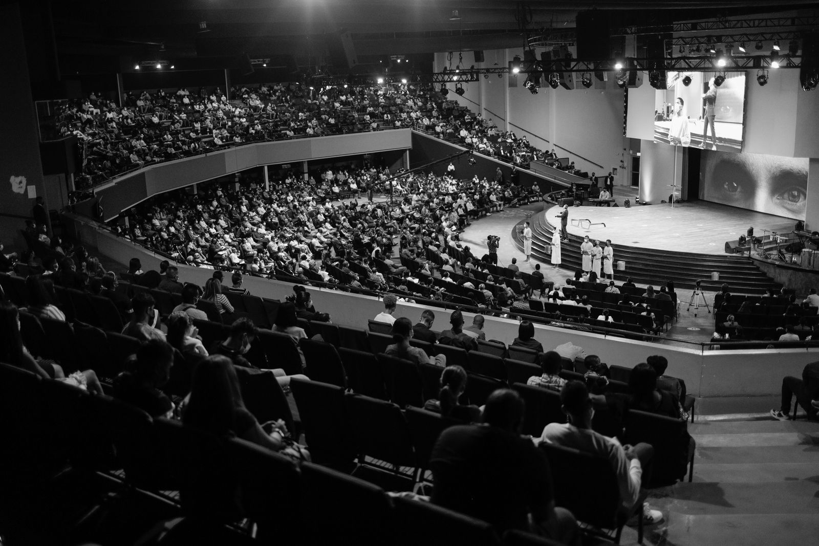 Foto em preto e branco de um auditório lotado visto do alto, com a plateia nas arquibancadas voltada para o palco iluminado e um grande telão ao fundo, evocando a era dos programas de auditório