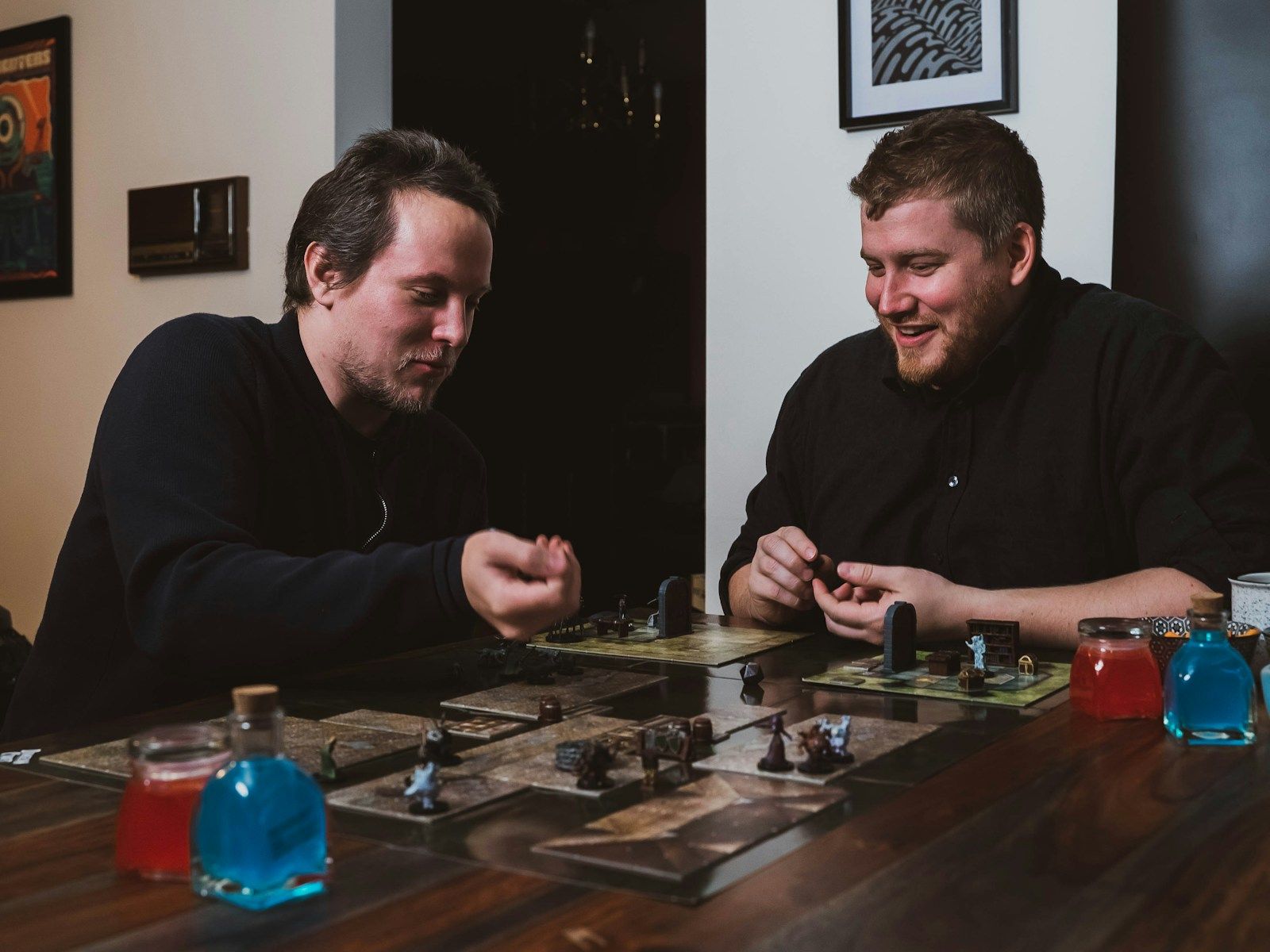 Dois amigos sorrindo em volta de uma mesa de RPG, movendo miniaturas sobre um mapa tático durante uma sessão de jogo