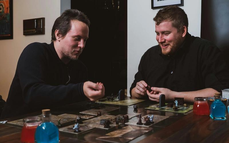 Dois amigos sorrindo em volta de uma mesa de RPG, movendo miniaturas sobre um mapa tático durante uma sessão de jogo
