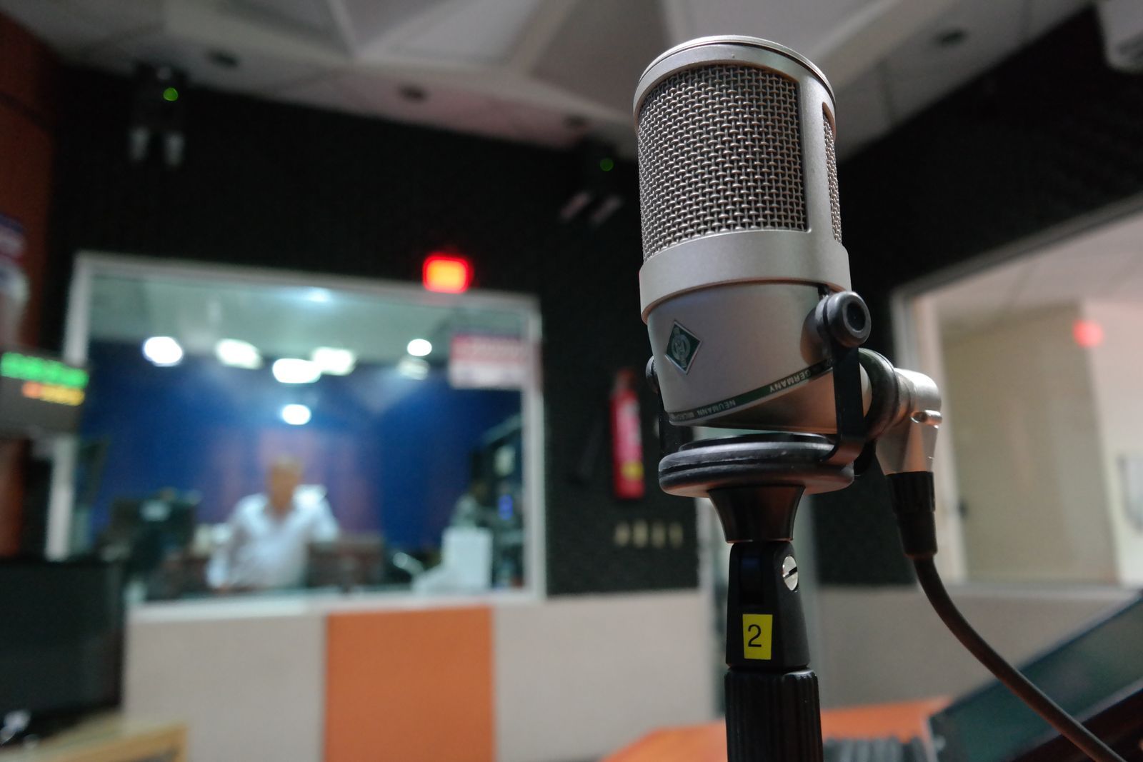 Microfone profissional de estúdio em primeiro plano dentro de uma emissora de rádio, com a cabine de transmissão e o operador desfocados ao fundo