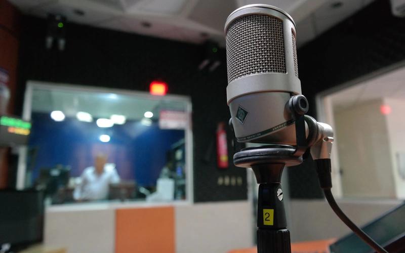 Microfone profissional de estúdio em primeiro plano dentro de uma emissora de rádio, com a cabine de transmissão e o operador desfocados ao fundo