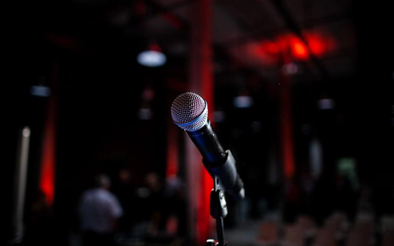 Microfone em um pedestal num palco escuro, iluminado por luzes vermelhas ao fundo, evocando a tensão do momento de falar diante do público.