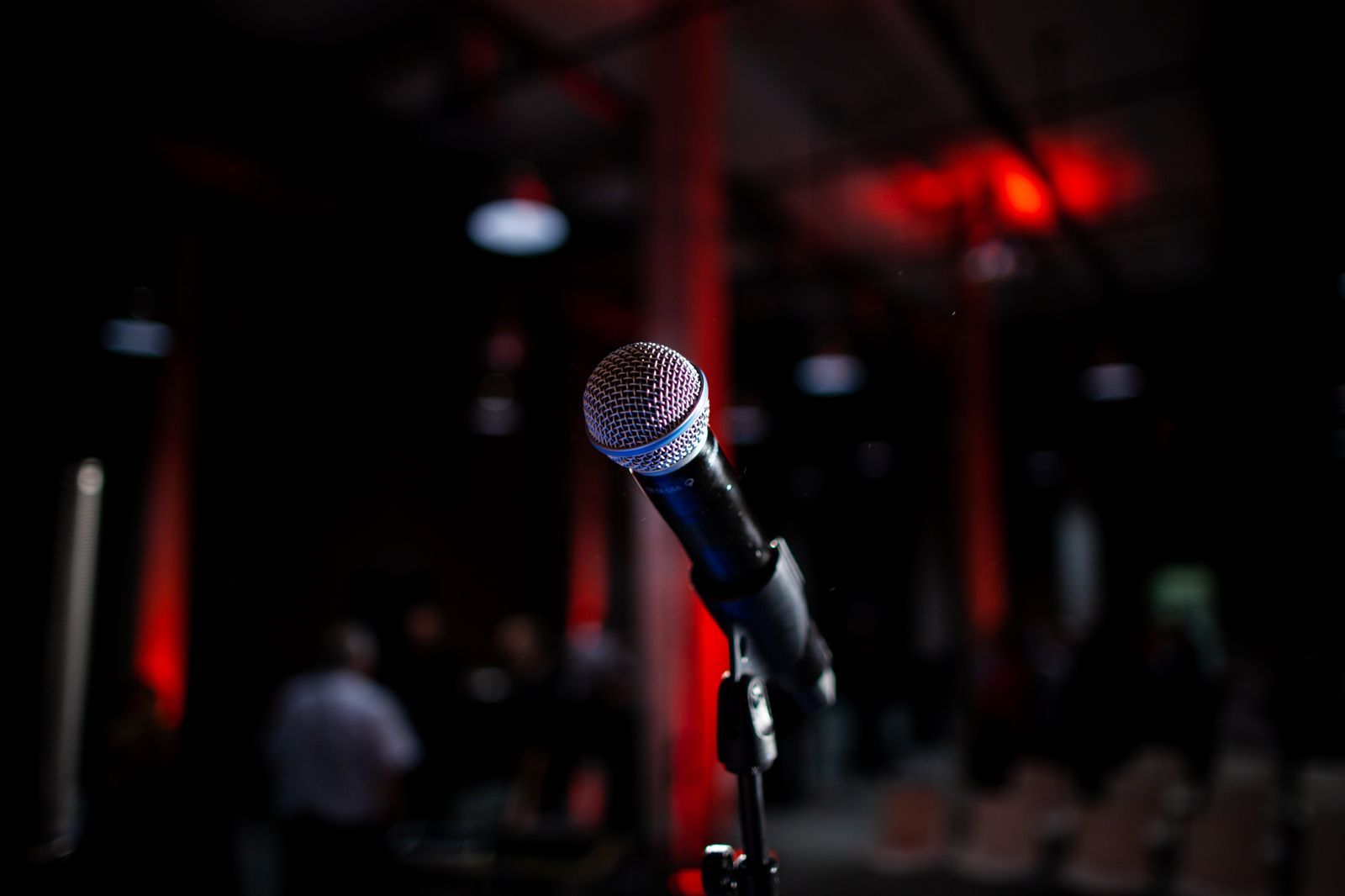 Microfone em um pedestal num palco escuro, iluminado por luzes vermelhas ao fundo, evocando a tensão do momento de falar diante do público.