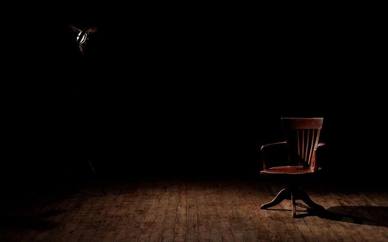 Cadeira de madeira vazia iluminada por um único spot de luz em um estúdio escuro, antes do início da programação