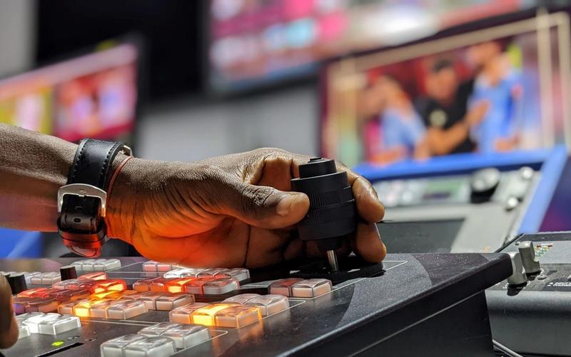 Mão operando os controles de uma mesa de transmissão em estúdio de TV, com telas desfocadas ao fundo mostrando o programa ao vivo