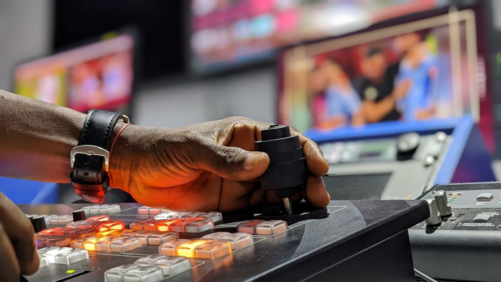 Mão operando os controles de uma mesa de transmissão em estúdio de TV, com telas desfocadas ao fundo mostrando o programa ao vivo