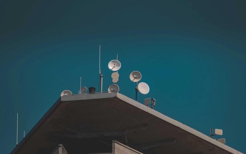 Antenas parabólicas de comunicação no topo de um prédio, contra um céu azul profundo, simbolizando a transmissão via satélite