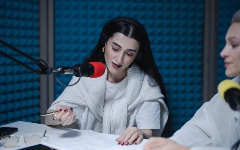 Locutora com fones de ouvido lê o roteiro apoiado na mesa enquanto fala ao microfone profissional dentro de um estúdio de rádio com isolamento acústico