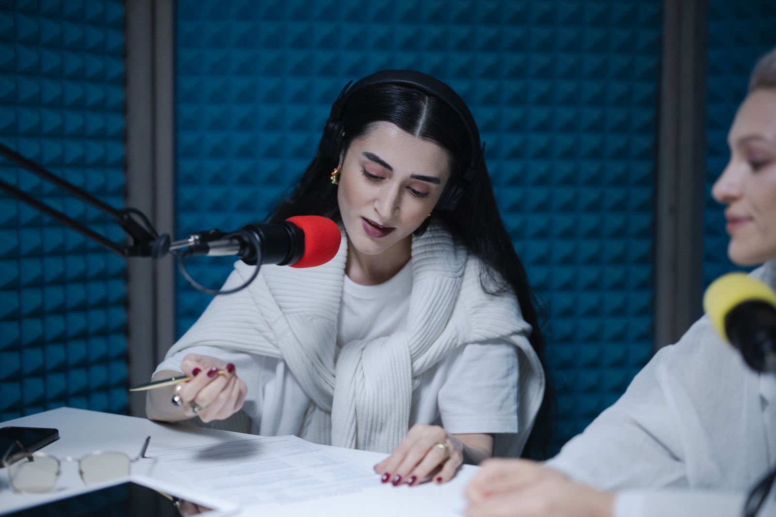 Locutora com fones de ouvido lê o roteiro apoiado na mesa enquanto fala ao microfone profissional dentro de um estúdio de rádio com isolamento acústico