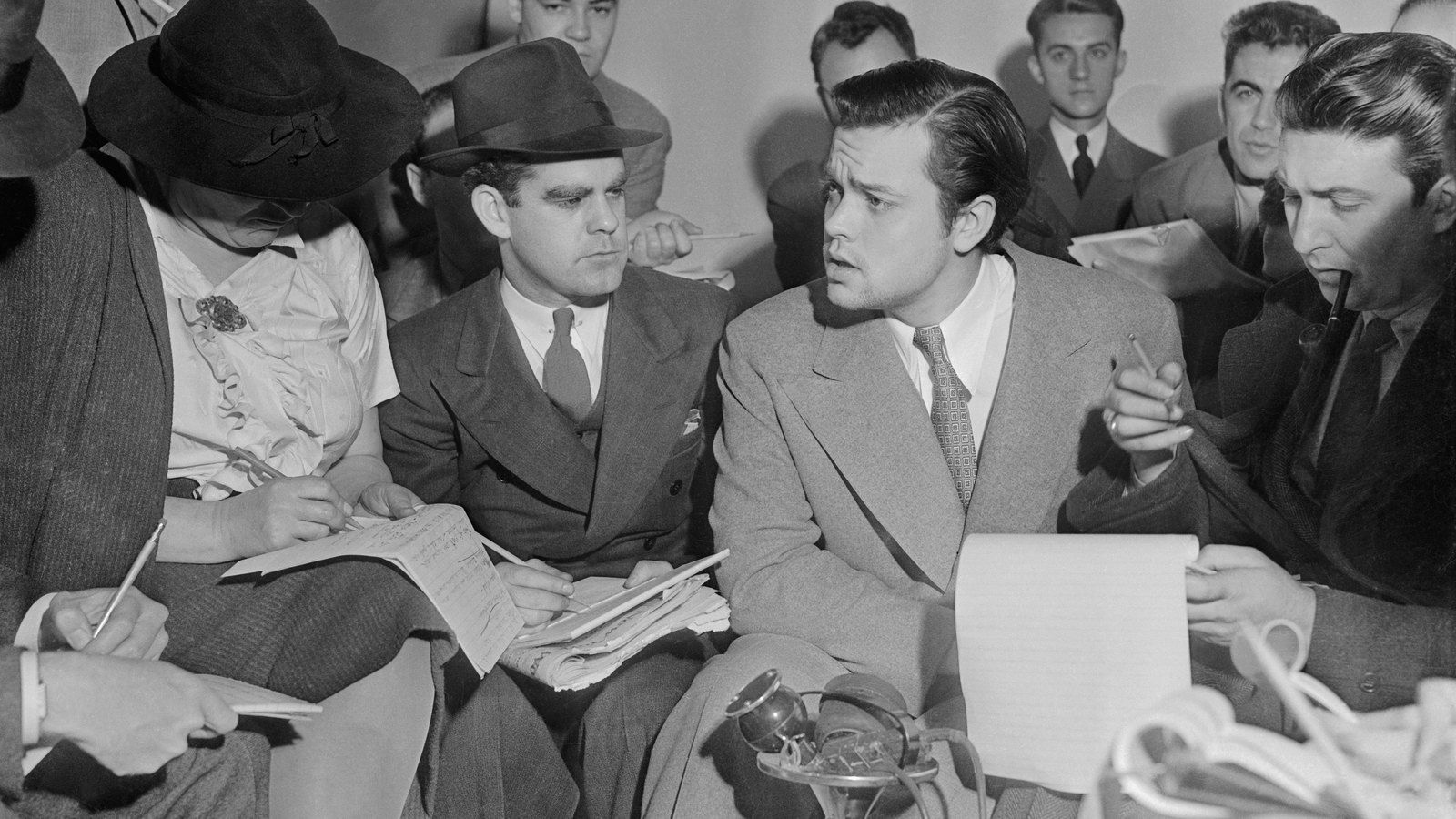 Orson Welles cercado de repórteres em 31 de outubro de 1938, um dia após a transmissão de A Guerra dos Mundos, com microfones à sua frente.