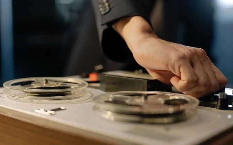 Close-up de uma mão ajustando os controles de um gravador de rolo (reel-to-reel) vintage, remetendo à gravação de jingles publicitários de rádio