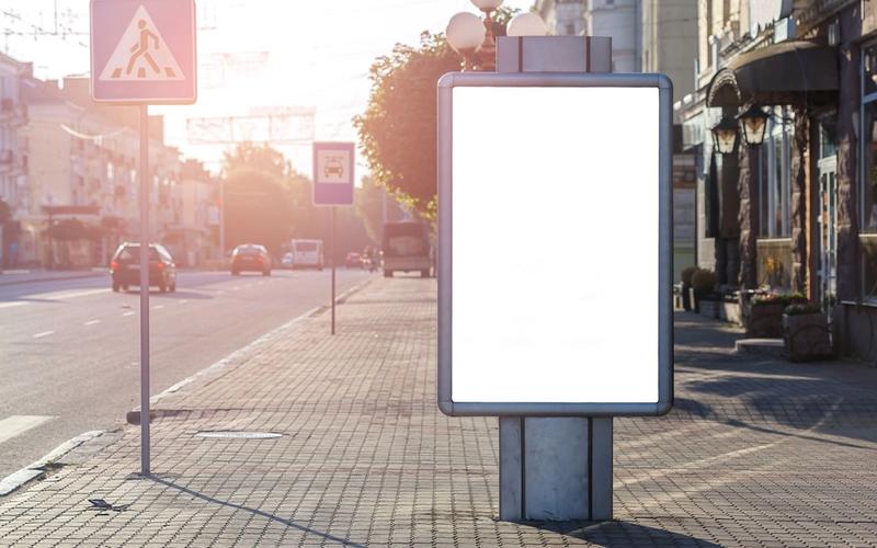 Painel de publicidade vazio em calçada de rua urbana sob luz dourada do fim de tarde, com carros e fachadas de lojas ao fundo