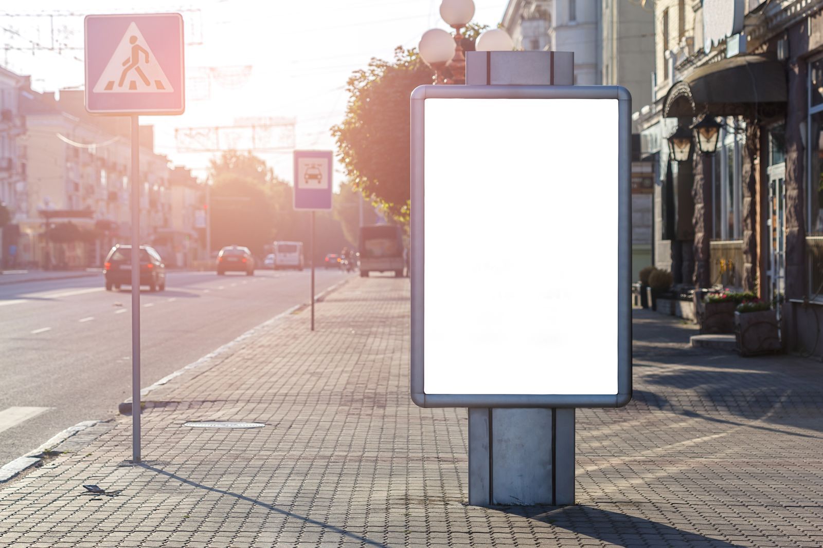 Painel de publicidade vazio em calçada de rua urbana sob luz dourada do fim de tarde, com carros e fachadas de lojas ao fundo