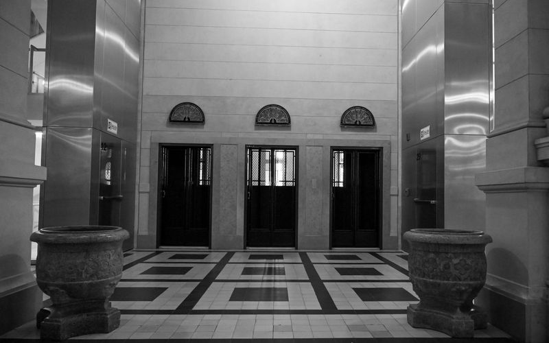 Lobby de elevadores estilo Art Déco em preto e branco, com três portas de grade metálica, arcos decorativos e piso geométrico em mármore