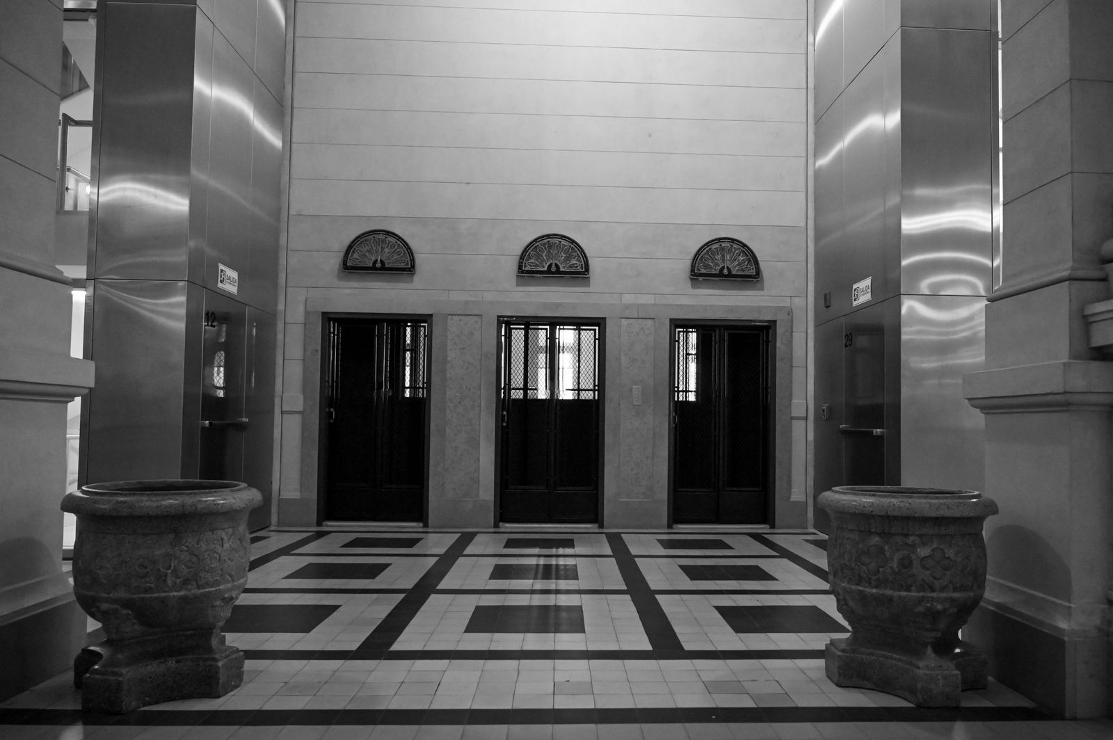 Lobby de elevadores estilo Art Déco em preto e branco, com três portas de grade metálica, arcos decorativos e piso geométrico em mármore