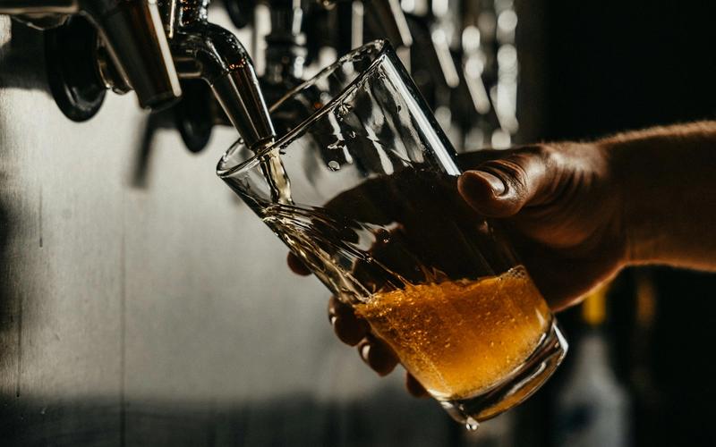 Chope dourado sendo servido em copo de vidro sob torneira de bar, com iluminação dramática e fundo escuro desfocado